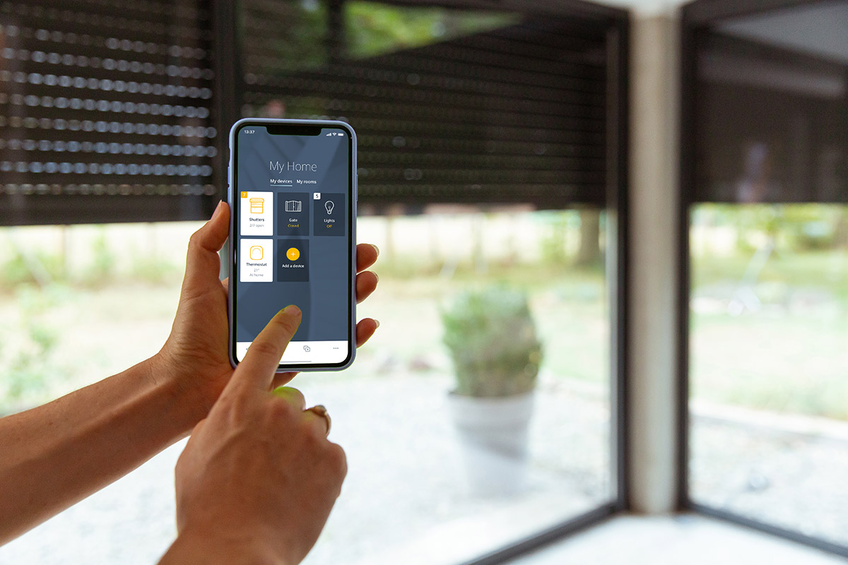 Mobilní telefon s aplikací pro ovládání rolet, venkovní rolety v pozadí.